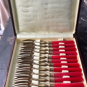Peut inclure: Un ensemble de fourchettes anciennes avec des manches rouges et des dents en métal doré, soigneusement disposées dans une boîte marron avec un intérieur crème. Les fourchettes sont probablement destinées au service ou à la dégustation.