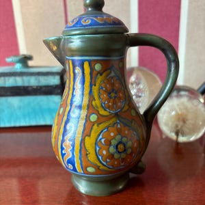 Może przedstawiać: Mały, zielony dzbanek ceramiczny z pokrywką. Dzbanek jest ozdobiony pomarańczowymi, żółtymi, niebieskimi i zielonymi wzorami kwiatowymi.
