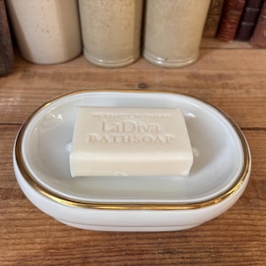 Può includere: Un portasapone in ceramica bianca con un bordo dorato, che presenta una saponetta bianca rettangolare con la scritta "La Diva Bathsoap" stampata su di essa.