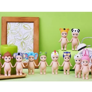 May include: A collection of 12 small, nude, plastic figurines wearing animal hats. The figurines are arranged in a row on a wooden surface. The hats are various colors and feature animals such as a monkey, koala, sloth, frog, owl, and tiger.