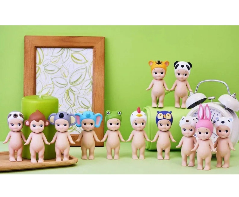 May include: A collection of 12 small, nude, plastic figurines wearing animal hats. The figurines are arranged in a row on a wooden surface. The hats are various colors and feature animals such as a monkey, koala, sloth, frog, owl, and tiger.