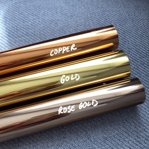 May include: Three cylindrical rolls of metallic foil arranged horizontally. The top roll is copper, the middle roll is gold, and the bottom roll is rose gold. Each roll features white handwritten text identifying the color as COPPER, GOLD, or ROSE GOLD.