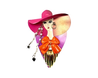 Puede incluir: Un broche de esmalte rosa y morado con una mujer con un gran sombrero rosa, un lazo naranja y joyas doradas.