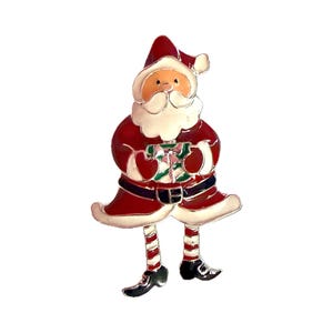 Puede incluir: Un broche de esmalte metálico con forma de Santa Claus que viste un traje rojo con bordes blancos, cinturón negro y gorro. Sostiene un regalo verde y blanco y tiene piernas largas con medias a rayas rojas y blancas y zapatos negros.