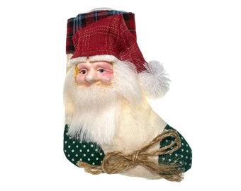 May include: A white stocking with a green and white polka dot pattern, a red and white plaid pattern, and a Santa Claus face with a white beard. The stocking has a brown twine bow.