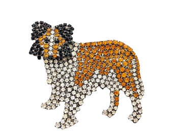 Puede incluir: Un broche de perro de cristal de roca. El perro es blanco con marcas naranjas y negras. Los cristales de roca son transparentes, naranjas y negros.