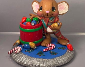 May include: A brown mouse figurine wearing a scarf, holding a Christmas light, and standing next to a red barrel filled with colorful lights. The figurine is on a blue base with Christmas trees and candy canes. The base has a silver glitter border.