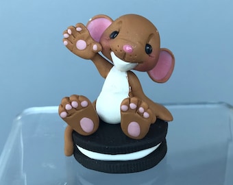 May include: A brown mouse figurine with pink ears and paws, waving its hand. The mouse is sitting on top of a black and white cookie. The figurine has a white belly and a pink nose. The mouse is smiling.