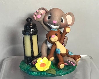 May include: A whimsical figurine featuring a gray mouse holding a brown teddy bear. The mouse has pink accents on its ears and paws. A black lantern and a yellow flower sit on a green base. The teddy bear has blue patches.