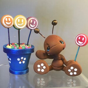 May include: A charming clay sculpture of a brown ant-like figure with blue eyes, holding a red smiley face lollipop. Next to it is a blue flower pot with smiley face lollipops and colourful candies. The pot is decorated with white flower accents.