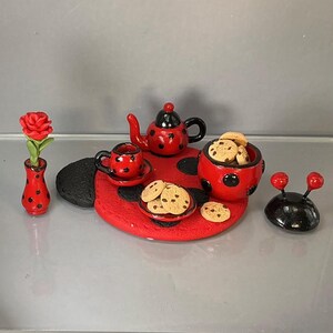 Könnte beinhalten: Ein Miniatur-Teeservice mit einer roten, schwarz gepunkteten Teekanne, einer passenden Tasse mit Untertasse, einer Schale mit Schokoladenkeksen, einer kleinen Vase mit einer roten Rose und einem schwarzen, marienkäferförmigen Untersetzer.