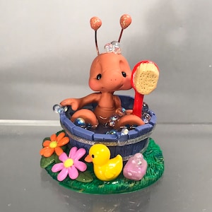 Puede incluir: Una figura caprichosa de una criatura marrón bañándose en una bañera azul con burbujas. Un cepillo rojo descansa a su lado. La base presenta flores, un pato amarillo y una piedra púrpura, todo sobre una base verde. La figura tiene dos antenas.
