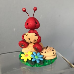 May include: A whimsical red ant figurine holding a chocolate chip cookie. The ant is sitting on a stack of cookies and a bed of green grass with yellow and blue flowers. The ant's antennae and feet have glitter accents.