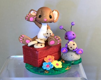 May include: A whimsical figurine featuring a brown mouse with pink accents, sitting on a red box and holding cookies. A purple ant with a cookie sits nearby. Colorful flowers and green grass surround the base.