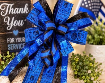 May include: A decorative bow made of blue and black ribbon with a metallic finish. The ribbon features printed text including "Blue," "Protect & Serve," "Hero," and "Brave," along with police badge icons. The bow has multiple loops and long tails.