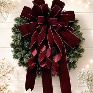 May include: A deep burgundy velvet ribbon bow with multiple loops and long, cascading tails attached to a circular green artificial pine wreath. The bow features a soft, plush texture and is centered on the foliage, creating a decorative display.