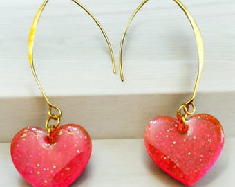 May include: A pair of gold dangle earrings with pink heart-shaped charms. The hearts have a glittery finish.
