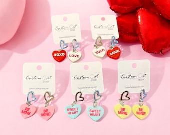 May include: Six pairs of heart-shaped earrings with different sayings. The earrings are on white cards with the words "Custom Cat Design" printed on them. The sayings on the earrings are "XOXO", "LOVE", "BE MINE", "SWEET HEART", and "BE MINE".