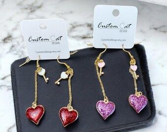 May include: A pair of dangle earrings with gold chains and heart-shaped charms. The charms are red, pink, and purple glitter with a key charm on each chain. The earrings are packaged in a black velvet box with a white card that reads "Custom Cat Design".