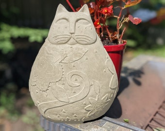 May include: A white concrete cat statue with a floral design. The cat is sitting on a stone surface with a red flower pot in the background.