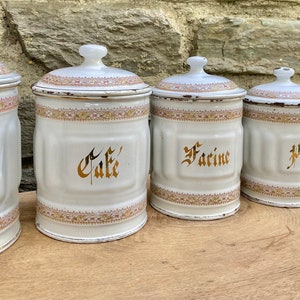 Peut inclure: Ensemble de six boîtes en émail blanc avec inscription dorée et bordure rose et dorée. Les boîtes sont étiquetées "Sucre", "Café", "Farine", "Pâtes", "Thé" et "Épices".