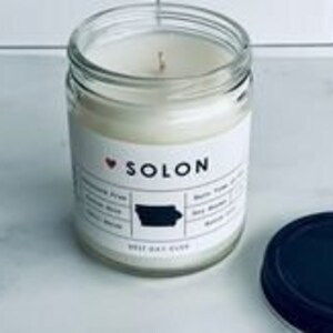 May include: A white candle in a clear glass jar with a black lid. The label on the jar says "SOLON" with a heart and a black outline of the state of Iowa. The candle is a soy wax blend with essential oils.