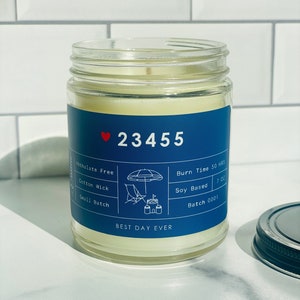 May include: A blue glass candle jar with a white label. The label features a red heart, the number 23455, and the text "BEST DAY EVER". The label also includes details about the candle, such as "Phthalate Free", "Cotton Wick", "Small Batch", "Burn Time 50 HRS", "Soy Based", and "Batch 0001".