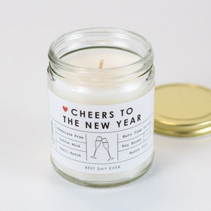 May include: A glass jar candle with a white label that reads "♥ CHEERS TO THE NEW YEAR". The label also includes details about the candle, such as "Paraben Free", "Cotton Wick", "Small Batch", "Burn Time 32 Hours", "Soy Based", and "Batch 2021". The jar has a gold lid.