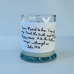 May include: A white candle in a clear glass jar with a white label. The label has black text that reads "Jesus said to him, "I am the way, and the truth, and the life. No one comes to the Father except through me." - John 14:6"