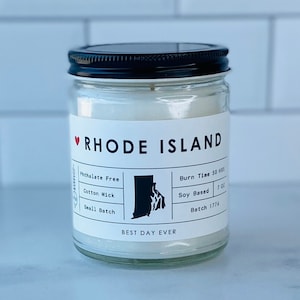 May include: A white candle in a clear glass jar with a black lid. The label says "Rhode Island" with a red heart and an outline of the state. The label also says "Phthalate Free", "Cotton Wick", "Small Batch", "Burn Time 50 hrs", "Soy Based 7 oz", and "Batch 1776". The bottom of the label says "Best Day Ever".