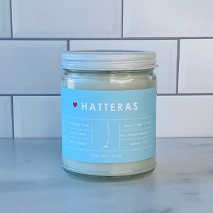 Op de afbeelding: Een witte kaars pot met een blauw label waarop "HATTERAS" staat met een rood hart. Het label vermeldt ook "BEST DAY EVER", "Soy Based Candle", "Batch 1670", "Cotton Wick", "Small Batch" en "Burn Time 50 hrs".