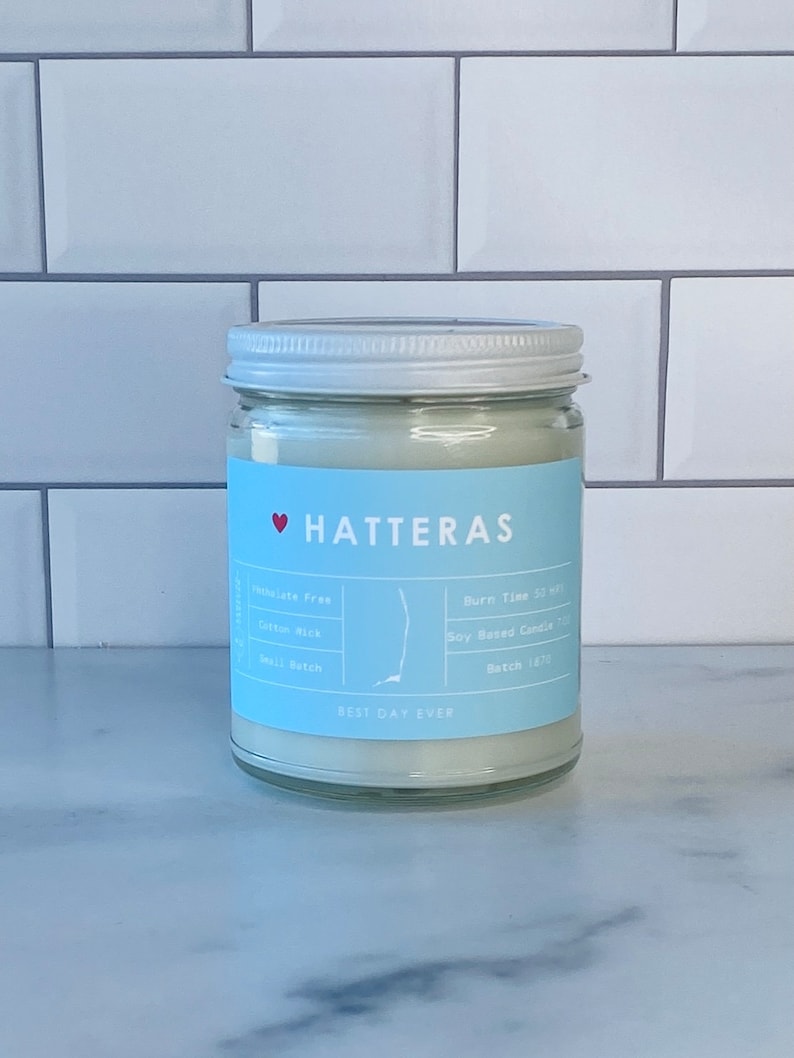 Op de afbeelding: Een witte kaars pot met een blauw label waarop "HATTERAS" staat met een rood hart. Het label vermeldt ook "BEST DAY EVER", "Soy Based Candle", "Batch 1670", "Cotton Wick", "Small Batch" en "Burn Time 50 hrs".