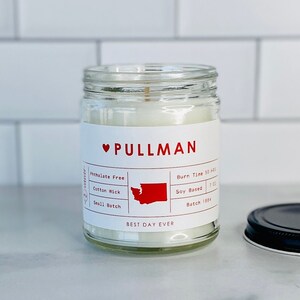 May include: A clear glass jar candle with a white label featuring the word "PULLMAN" in red. The label also includes the text "Phthalate Free", "Cotton Wick", and "Small Batch". The candle has a burn time of 50 hours and is soy-based.