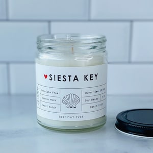 May include: A white candle in a clear glass jar with a black and white label that says "SIESTA KEY" and "BEST DAY EVER". The label also includes details about the candle, such as "Phthalate Free", "Cotton Wick", "Small Batch", "Burn Time 50 HRS", "Soy Based 7 OZ", and "Batch 1952".