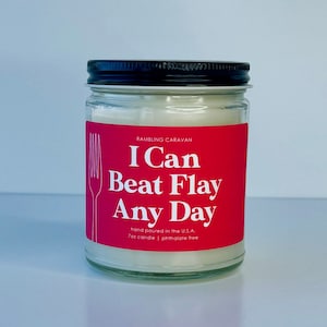 May include: A 7-ounce candle in a clear glass jar with a black lid. The label is pink with white text that reads "RAMBLING CARAVAN I Can Beat Flay Any Day hand poured in the U.S.A. 7oz candle | phthalate free".