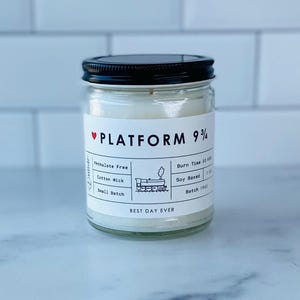 May include: A white candle in a clear glass jar with a black lid. The label reads "PLATFORM 9 3/4" with a red heart and a small train illustration. The label also lists details like "Phthalate Free", "Cotton Wick", "Small Batch", "Burn Time 50 hrs", "Soy Based", and "Batch 1961". The bottom of the label reads "BEST DAY EVER".
