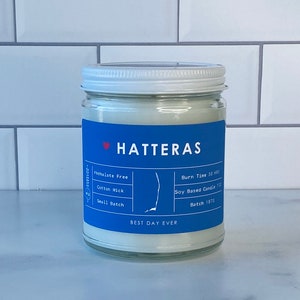 Op de afbeelding: Een witte kaars pot met een blauw label waarop "HATTERAS" en "BEST DAY EVER" staat. Het label bevat ook details over de kaars, zoals "Phthalate Free", "Cotton Wick", "Small Batch", "Burn Time 50 HRS", "Soy Based Candle 700" en "Batch 1870".
