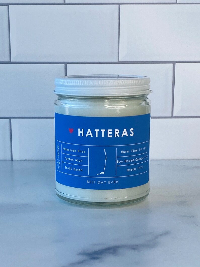Op de afbeelding: Een witte kaars pot met een blauw label waarop "HATTERAS" en "BEST DAY EVER" staat. Het label bevat ook details over de kaars, zoals "Phthalate Free", "Cotton Wick", "Small Batch", "Burn Time 50 HRS", "Soy Based Candle 700" en "Batch 1870".