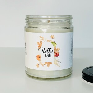 May include: A white candle in a clear glass jar with a label that says "Hello Fall". The label features a watercolor illustration of fall leaves, pumpkins, and gourds.