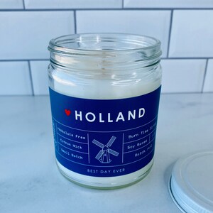 May include: A blue and white candle jar with a red heart and the word "HOLLAND" on the label. The label also includes the text "Phthalate Free", "Cotton Wick", "Small Batch", "Burn Time 50", "Soy Based", and "Batch 1400". The candle jar is sitting on a white surface.