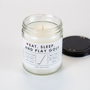 May include: A white soy candle in a glass jar with a black lid. The label reads "♥EAT, SLEEP, AND PLAY GOLF" with a golf club graphic. The candle is soy-based and has a cotton wick.