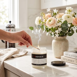 May include: A clear glass jar candle with a black label featuring the text BOUGIE, AF, and BEST DAY EVER. The candle is being lit with a match, producing a thin trail of smoke. The label also includes details like Phthalate Free and Soy Based.
