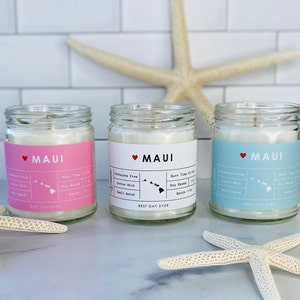 May include: Three glass candle jars containing white soy wax and featuring pink, white, and blue labels. Each label displays the text "MAUI" with a red heart and an outline of the Hawaiian island of Maui. The text "Best Day Ever" is printed on the bottom of each label.