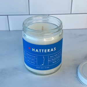 Op de afbeelding: Een blauwe en witte kaars pot met de tekst "HATTERAS" en "BEST DAY EVER". De kaars is op basis van soja en heeft een katoenen lont. De pot is versierd met een kleine omtrek van de staat North Carolina. De kaars is ftalaatvrij en heeft een brandtijd van 50 uur. Batchnummer 1870.