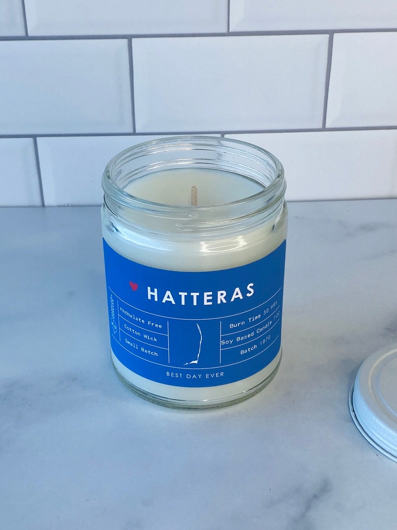 Op de afbeelding: Een blauwe en witte kaars pot met de tekst "HATTERAS" en "BEST DAY EVER". De kaars is op basis van soja en heeft een katoenen lont. De pot is versierd met een kleine omtrek van de staat North Carolina. De kaars is ftalaatvrij en heeft een brandtijd van 50 uur. Batchnummer 1870.