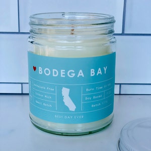 Könnte beinhalten: Ein blaues und weißes Kerzengefäß mit dem Text "♡ BODEGA BAY" und "BEST DAY EVER". Die Kerze ist soja-basiert und hat einen Baumwolldocht. Das Gefäß enthält auch den Text "Phthalate Free", "Burn Time 50 HRS", "Soy Based", "Small Batch", und "Batch 1775".