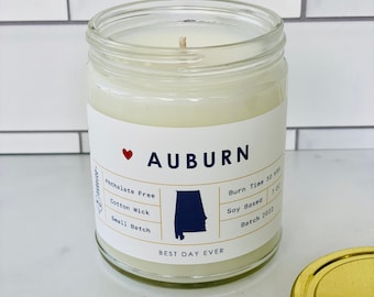 Op de afbeelding: Een witte kaars pot met een gouden deksel. Het etiket zegt "AUBURN" met een rood hart en een omtrek van de staat Alabama. Het etiket zegt ook "Ftalatenvrij", "Katoenen lont", "Kleine batch", "Brandtijd 50 uur", "Soja gebaseerd" en "Batch 2022". Het etiket zegt ook "Best Day Ever".
