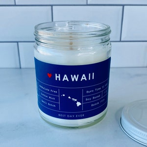 May include: A blue and white candle jar with a red heart and the text "HAWAII" and "BEST DAY EVER". The jar is labelled with the text "Paraben Free", "Cotton Wick", "Small Batch", "Burn Time 50 HRS", "Soy Based", and "Batch 1959".