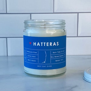 Op de afbeelding: Een blauwe en witte kaars pot met de tekst "HATTERAS" en "BEST DAY EVER". De kaars is op basis van soja en heeft een brandtijd van 50 uur. De pot is gelabeld met "Ftalatenvrij", "Katoenen lont" en "Kleine batch".