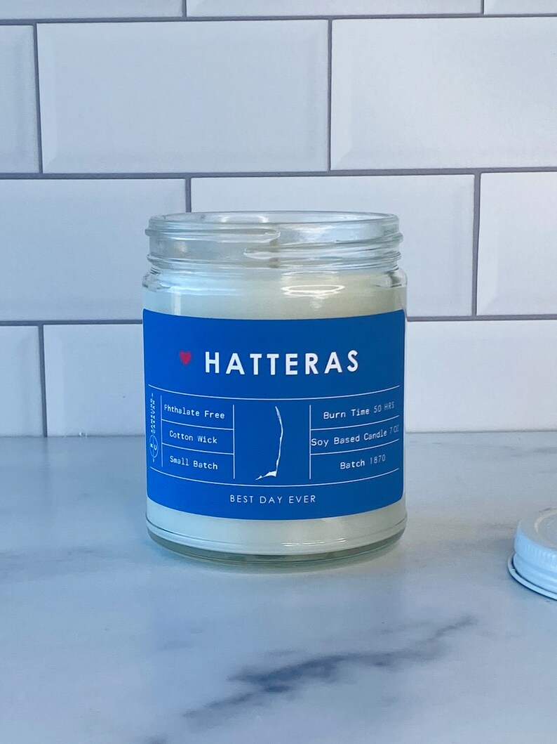 Op de afbeelding: Een blauwe en witte kaars pot met de tekst "HATTERAS" en "BEST DAY EVER". De kaars is op basis van soja en heeft een brandtijd van 50 uur. De pot is gelabeld met "Ftalatenvrij", "Katoenen lont" en "Kleine batch".